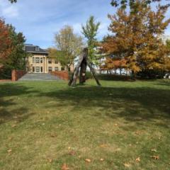 A sculpture on a lawn with surrounding fall foliage and a building in the background